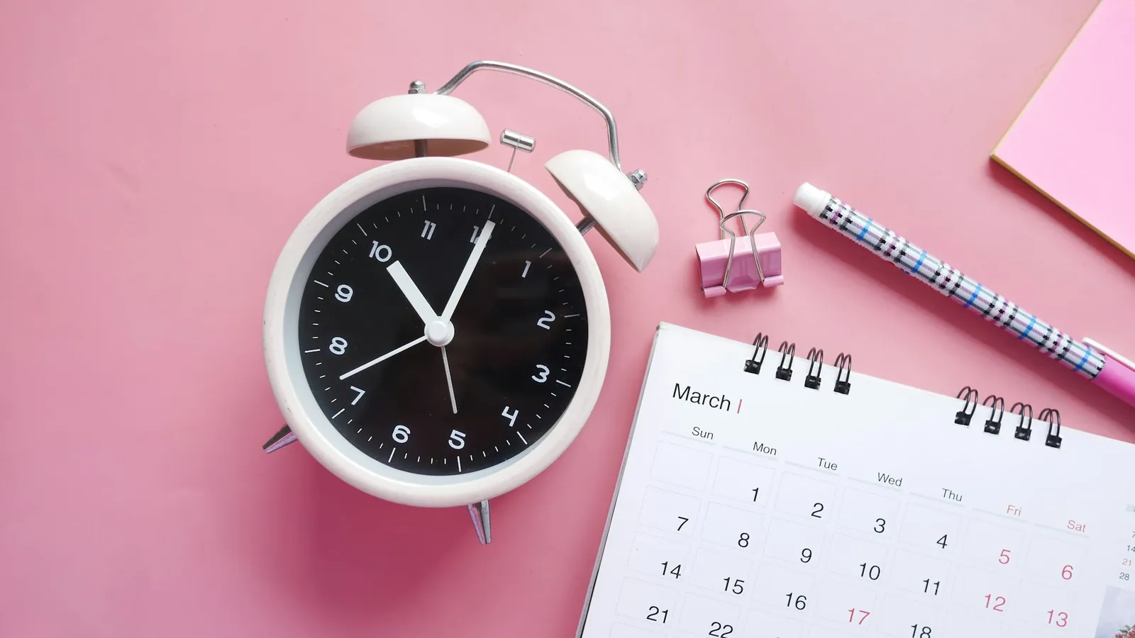 An alarm clock beside a desk calendar open to March, on a pink desk